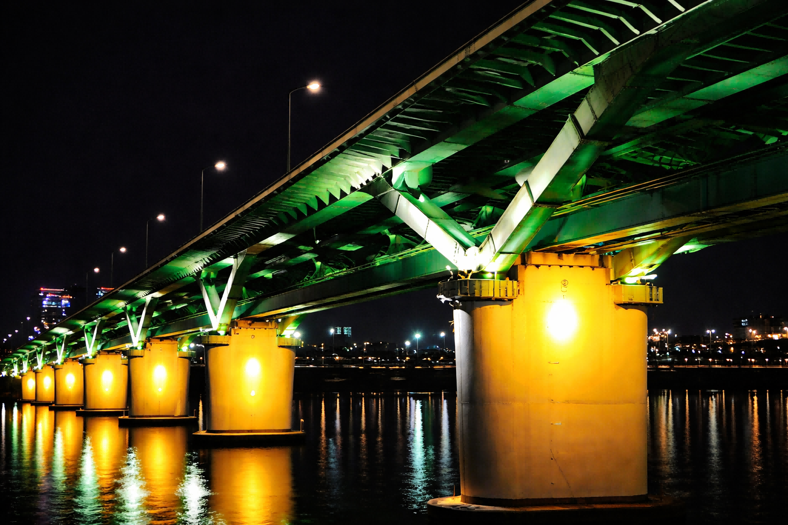Cấu trúc cầu hiện đại được chiếu sáng bởi đèn pha LED màu xanh lá cây và vàng vào ban đêm, cho thấy sự phân bố ánh sáng đồng đều và phản chiếu trên nước.